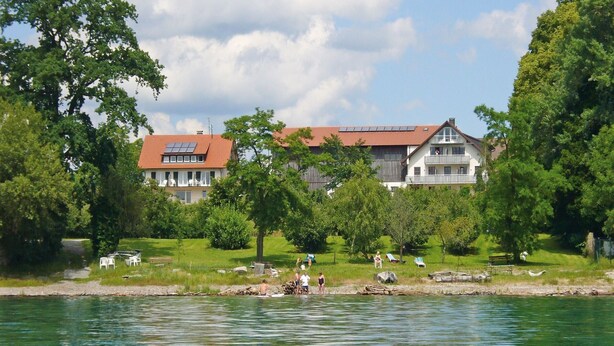 A lakeside view with a house featuring a red roof and solar panels, surrounded by greenery and trees.