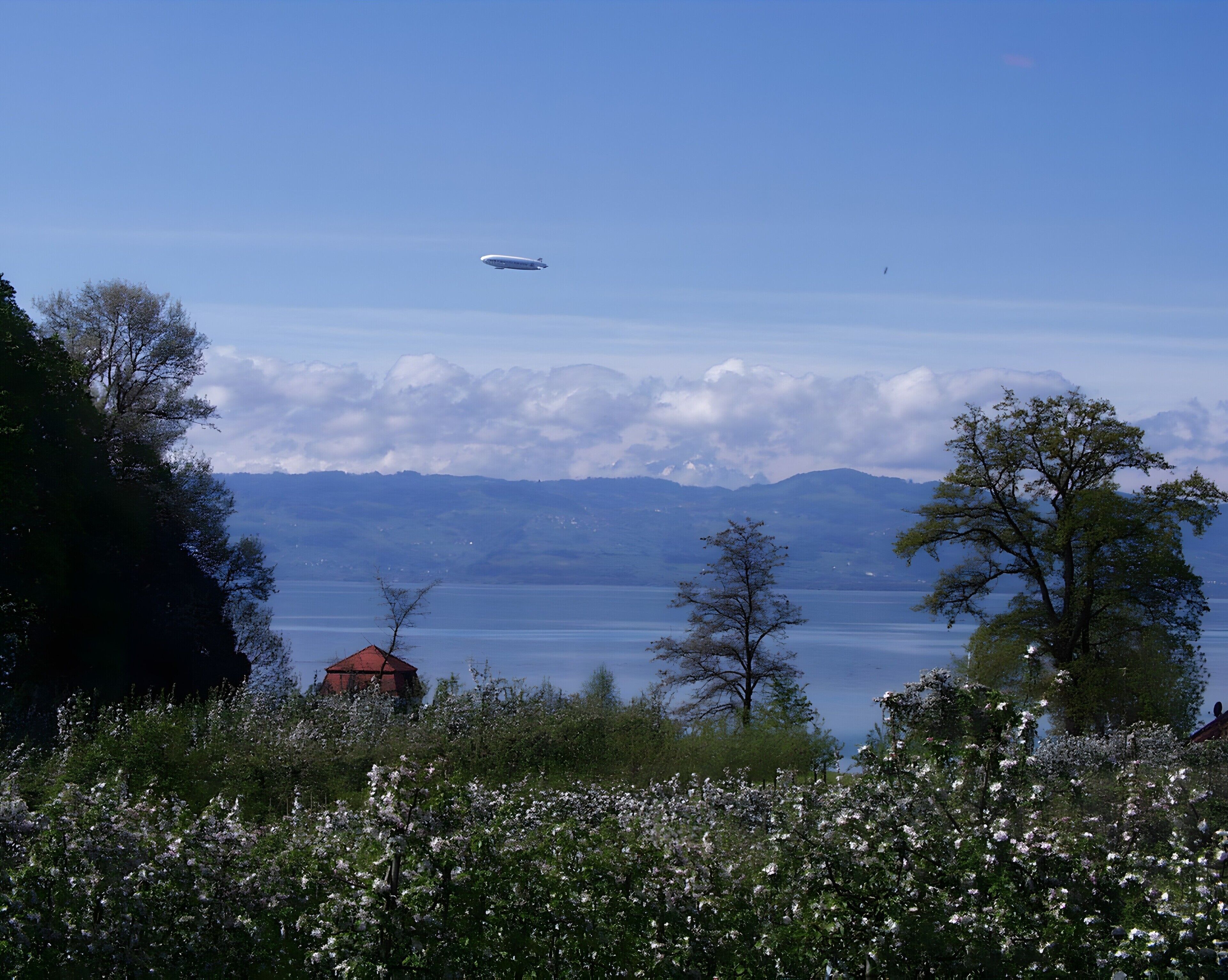 A scenic view of a lake with mountains in the background, a red-roofed building, and a white airplane in the sky.