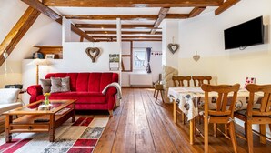 Una sala de estar acogedora con un sofá rojo, una mesa de comedor de madera y sillas, vigas de madera en el techo y un televisor montado en la pared.