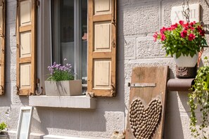 A building facade with a window with wooden shutters, a heart-shaped decoration, and a potted plant with flowers.