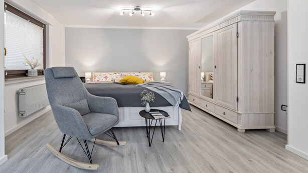 Ein Schlafzimmer mit einem Bett, einem Schaukelstuhl, einem Kleiderschrank und einem kleinen Tisch mit einer Vase.