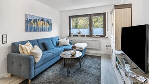 Ein modernes Wohnzimmer mit einem blauen Sofa, einem Couchtisch, einem Flachbildfernseher und einem großen Fenster mit Jalousien.