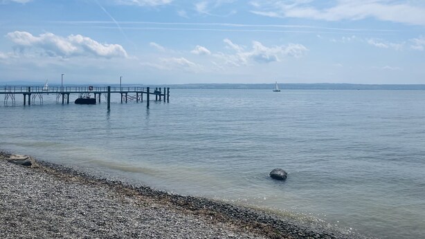 Una orilla tranquila con un muelle que se adentra en el agua, un velero a lo lejos y una playa de guijarros en primer plano.