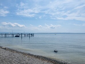 Una tranquilla riva con un molo che si protende sull'acqua, una barca a vela all'orizzonte e una spiaggia di ciottoli in primo piano.