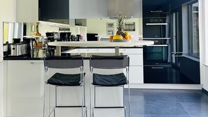 Una cocina moderna con una isla central, dos taburetes de bar, un frutero y un ventanal grande.