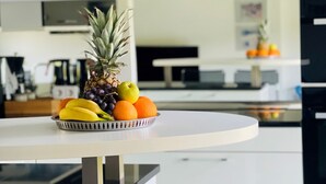 Una cocina moderna con un frutero sobre una mesa blanca, una cafetera y un refrigerador al fondo.
