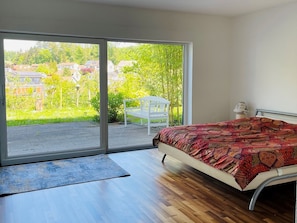 Camera da letto con un letto, una panca e vista su giardino e edifici attraverso una porta scorrevole in vetro.
