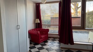 Ein Zimmer mit kariertem Boden, einem roten Sofa, einem weißen Kleiderschrank, einem Fenster mit Jalousien und einer Lampe.