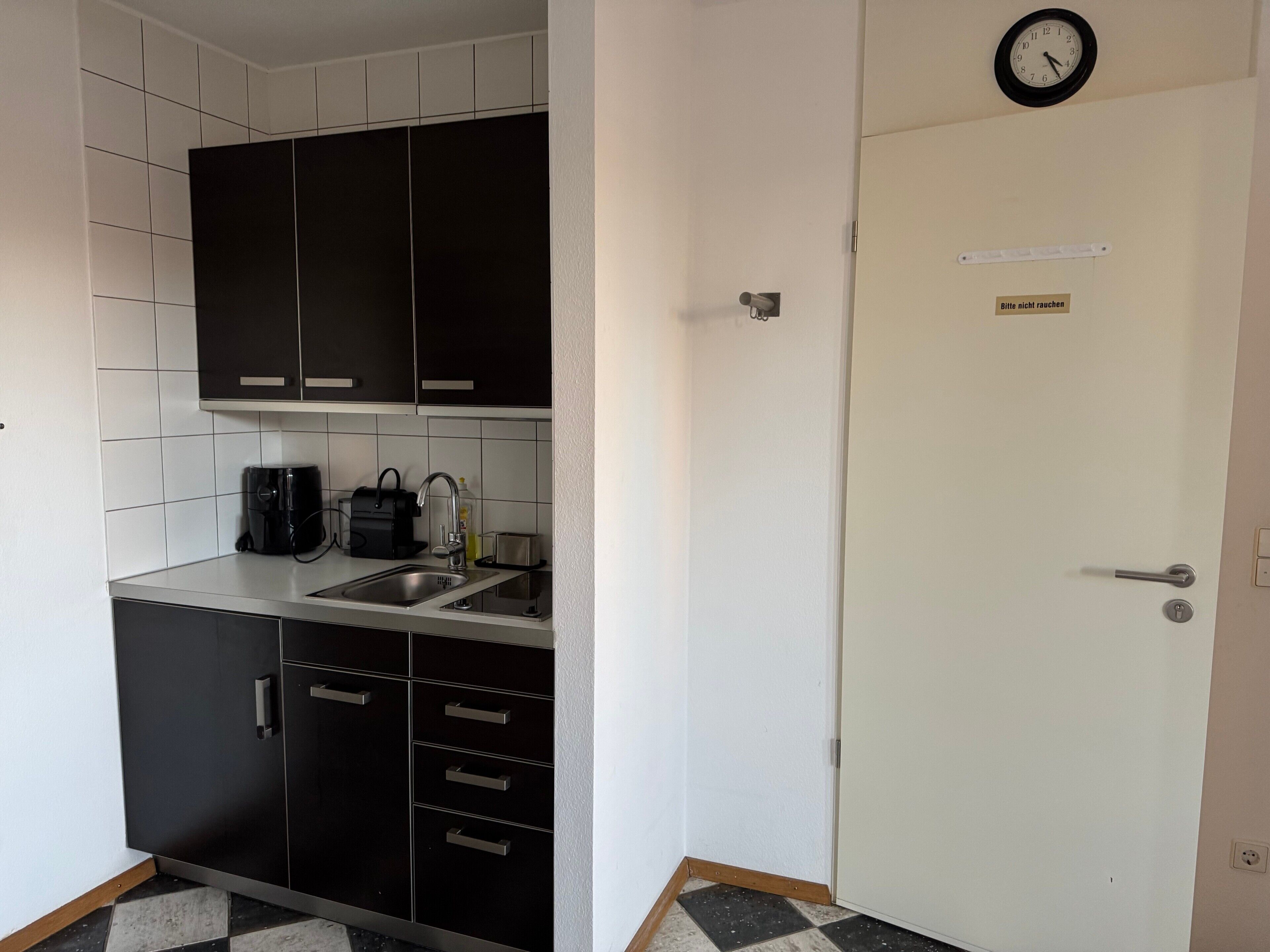 A small kitchenette with a sink, a coffee maker, and a toaster. There is a door with a 'Please do not smoke' sign above it.
