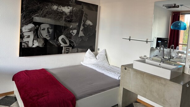 Ein modernes Schlafzimmer mit einem großen abstrakten Gemälde, einem Bett mit roter Tagesdecke, einer schlichten Kommode und einem großen Spiegel.