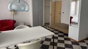 Una habitación con una mesa blanca, un sofá rojo, suelo a cuadros, un armario gris y un espejo.