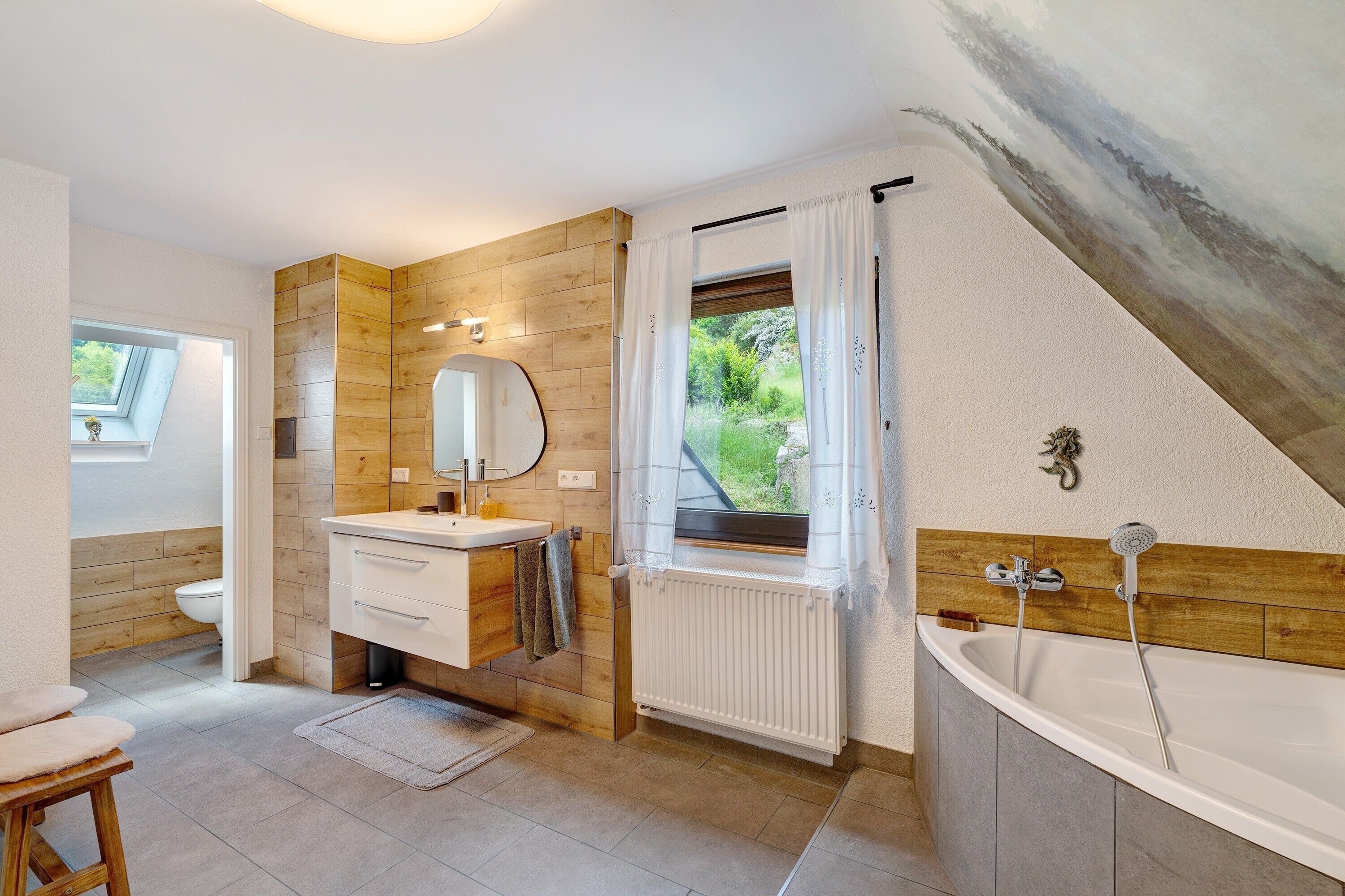 Un baño con pared de madera, un lavabo blanco, un espejo, una ventana con cortinas y una bañera.
