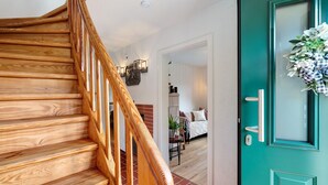 Un escalier en bois mène à une pièce dotée d’une porte de couleur turquoise, d’un lit et d’une petite table sur laquelle est posée une plante.