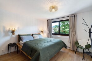 Un dormitorio con cama, mesitas de noche, una ventana con cortinas, una silla y una lámpara fijada a la pared.
