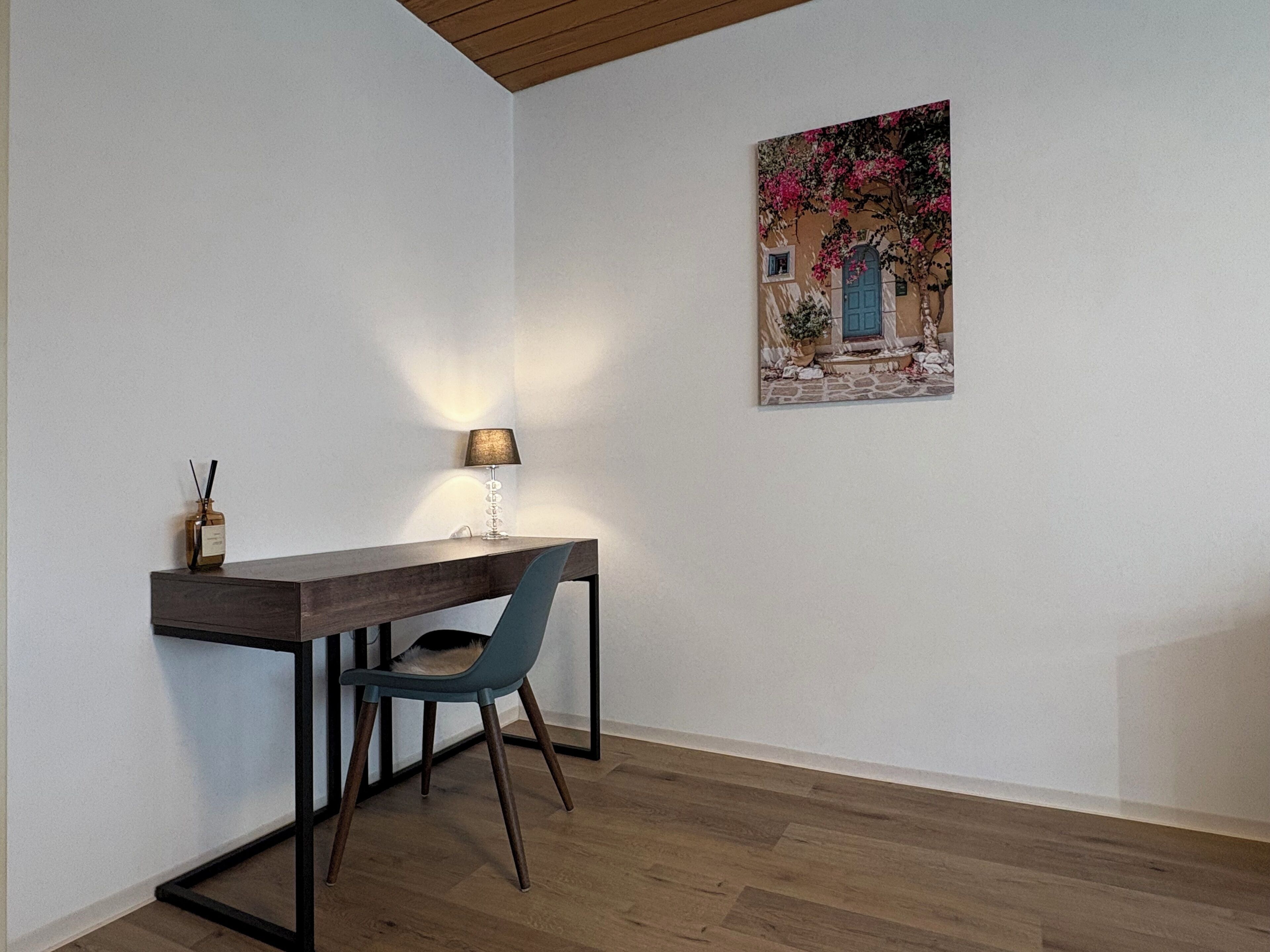 Ein Zimmer mit einem Holztisch, einem Stuhl, einer Lampe und einem Bild von einem Gebäude mit Blumen.