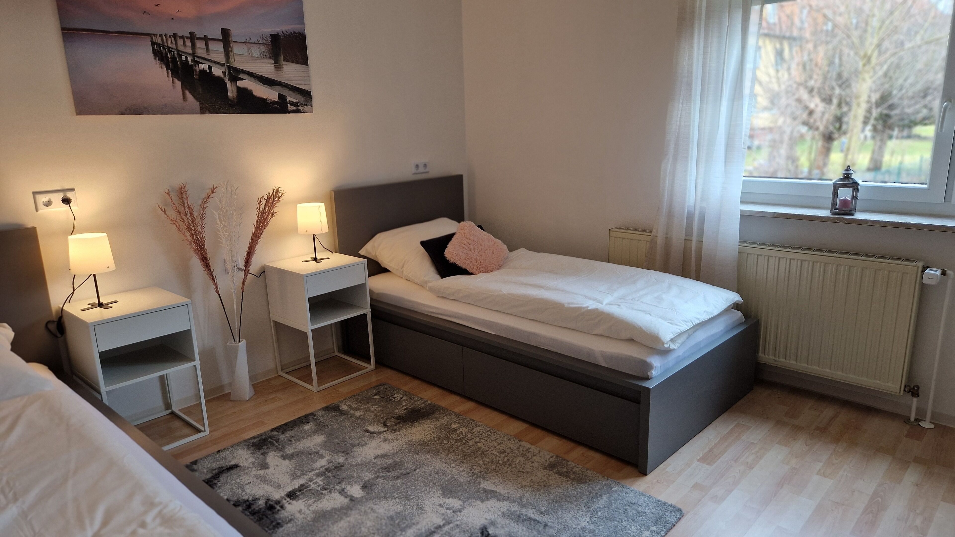 A bedroom with a bed, bedside tables, a nightstand, a radiator, and a window with a view of trees.