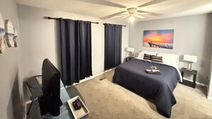 A hotel room with a bed, a desk, a chair, a ceiling fan, and a painting of a sunset over a pier. - Cozy Retreat w/ LED & Balcony (Charlotte)