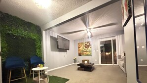 A living room with a ceiling fan, a flat-screen TV, a green wall, and a sliding glass door leading to a balcony. - Cozy Retreat w/ LED & Balcony (Charlotte)
