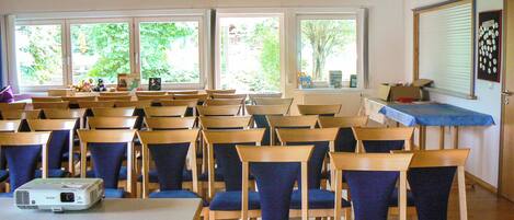 A classroom with wooden desks and chairs, a projector, and large windows with a view of greenery outside.