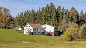 A large white building with multiple windows and a sloped roof, surrounded by a grassy field and dense forest. - Berger's Bergsicht by edita (Scheidegg)