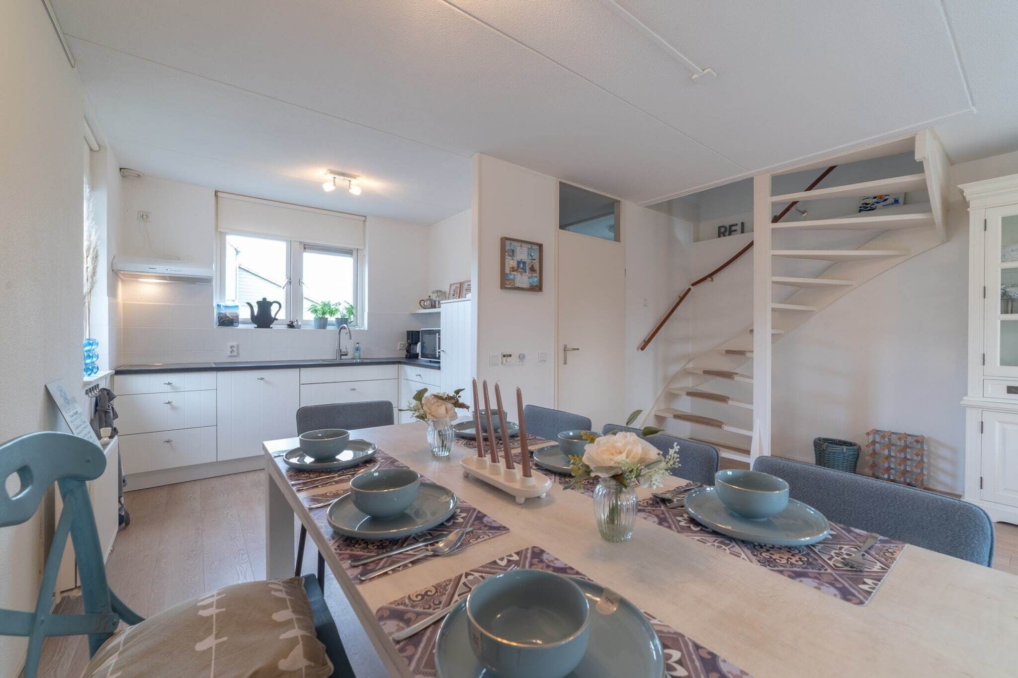 A modern kitchen with white cabinets, a dining table set with blue plates and bowls, and a staircase with a wooden railing.
