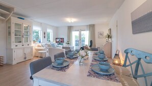 Et spiseområde med et langt bord dækket til et måltid, et hvidt skab med glasdøre og en sofa i baggrunden.