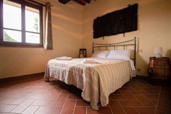 A bedroom with a bed, a window, a wooden barrel, and a wall-mounted shelf.