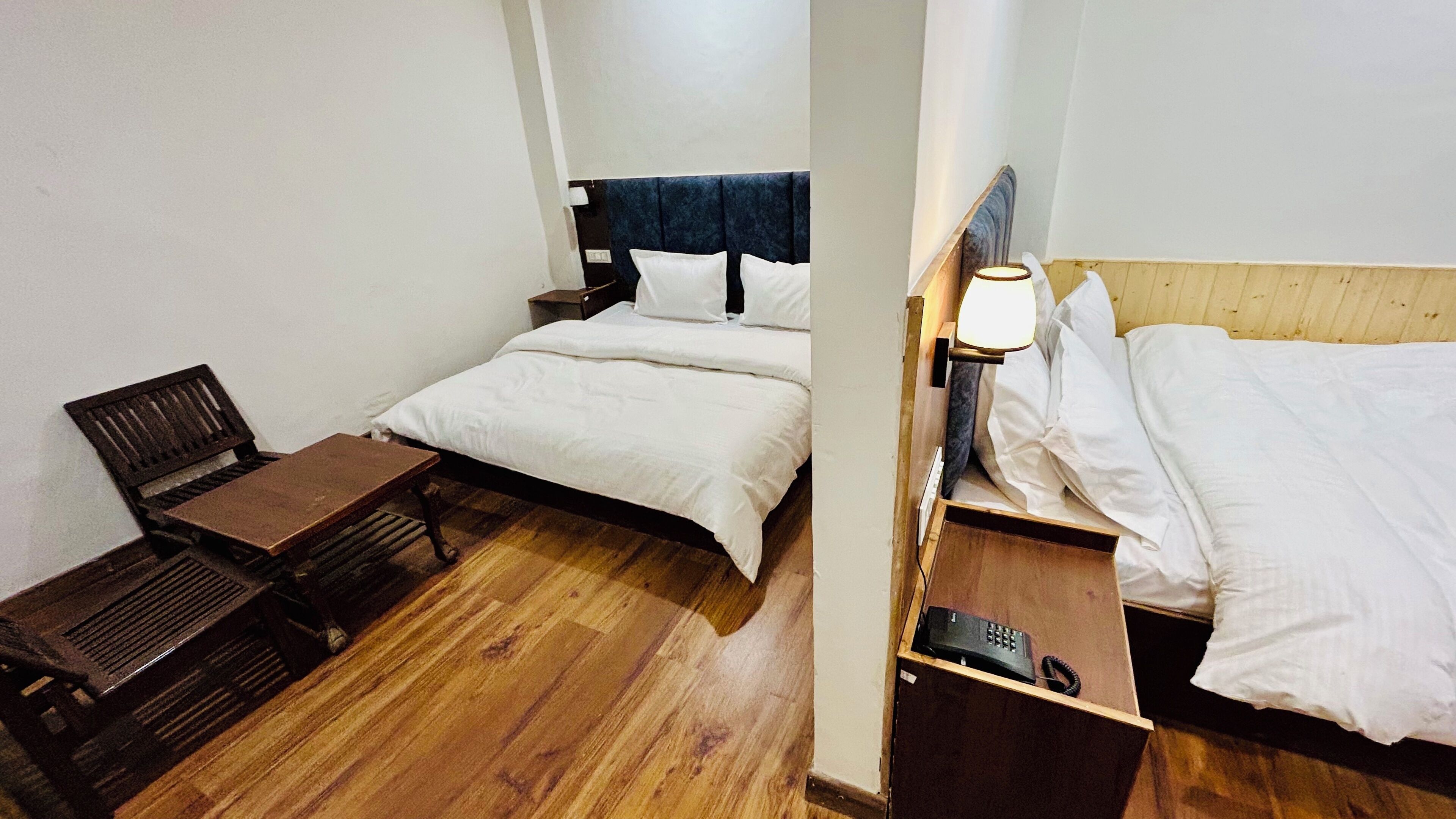 A small, compact hotel room with a single bed, a wooden nightstand with a lamp, a small table, and a chair.