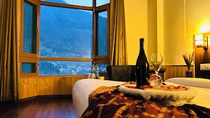 Une chambre d’hôtel dotée d’une grande fenêtre donnant sur un paysage montagneux. On y trouve un lit recouvert d’une couette blanche et un plateau décoratif avec une bouteille et un verre.