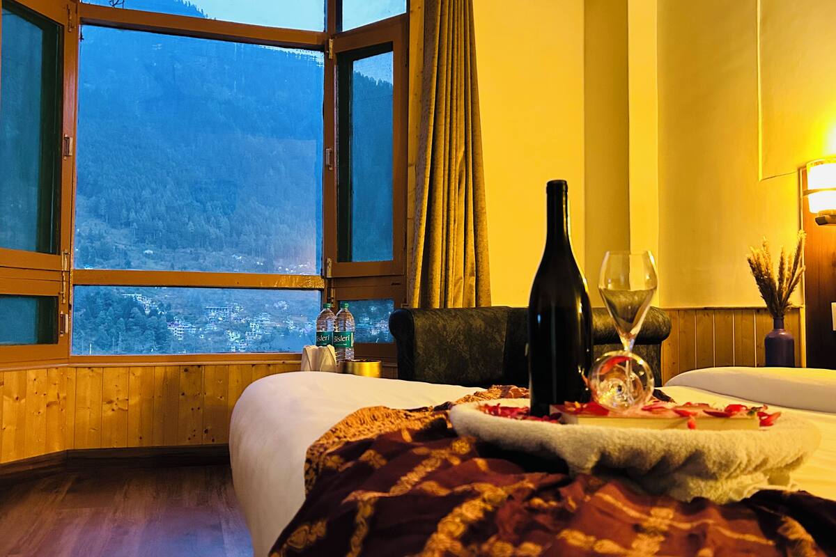 A hotel room with a large window offering a view of a mountainous landscape. There is a bed with a white comforter and a decorative tray with a bottle and glass.