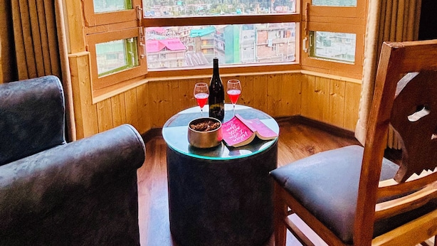 Une chambre d’angle dotée d’une grande baie vitrée donnant sur les montagnes. On y trouve une table ronde avec une bouteille et des verres, ainsi que deux chaises.