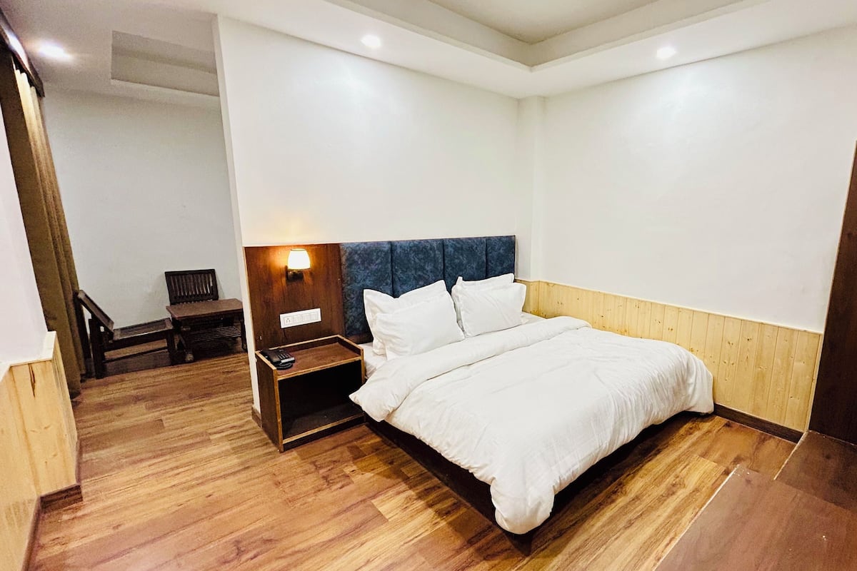 A hotel room with a wooden floor, a bed with white bedding, a nightstand with a lamp, and a wooden door.