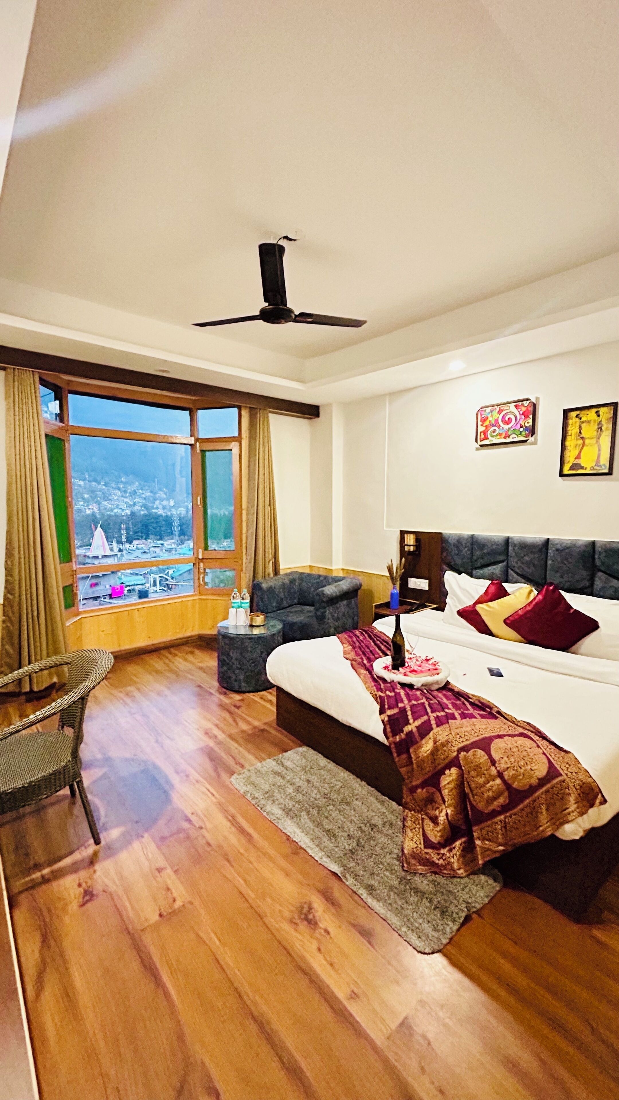 Habitación de hotel con una cama grande, ventilador de techo, vista panorámica de la ciudad, una mesita redonda y una silla de mimbre.