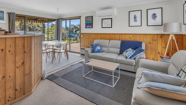 A modern living room with a wooden accent wall, a grey sofa, a coffee table, and a dining area with a white table and chairs.