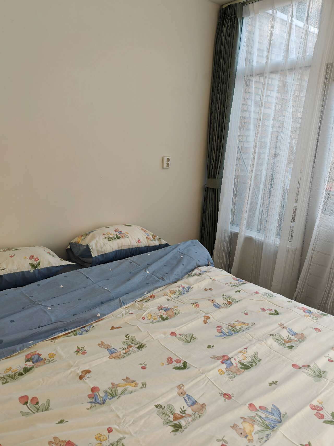 Ein Schlafzimmer mit einem Bett, floralem Bettbezug, einem Fenster mit Vorhängen und einer Steckdose.