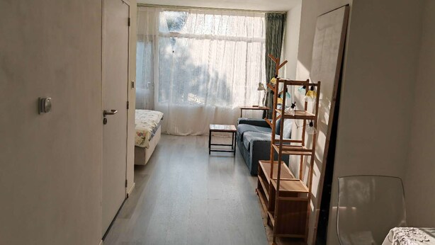 A narrow room with a bed, a sofa, a small table, and a wooden shelf unit. - Studio Breda Modern Furnishing space with a comfortable, 4min drive station. (Breda)
