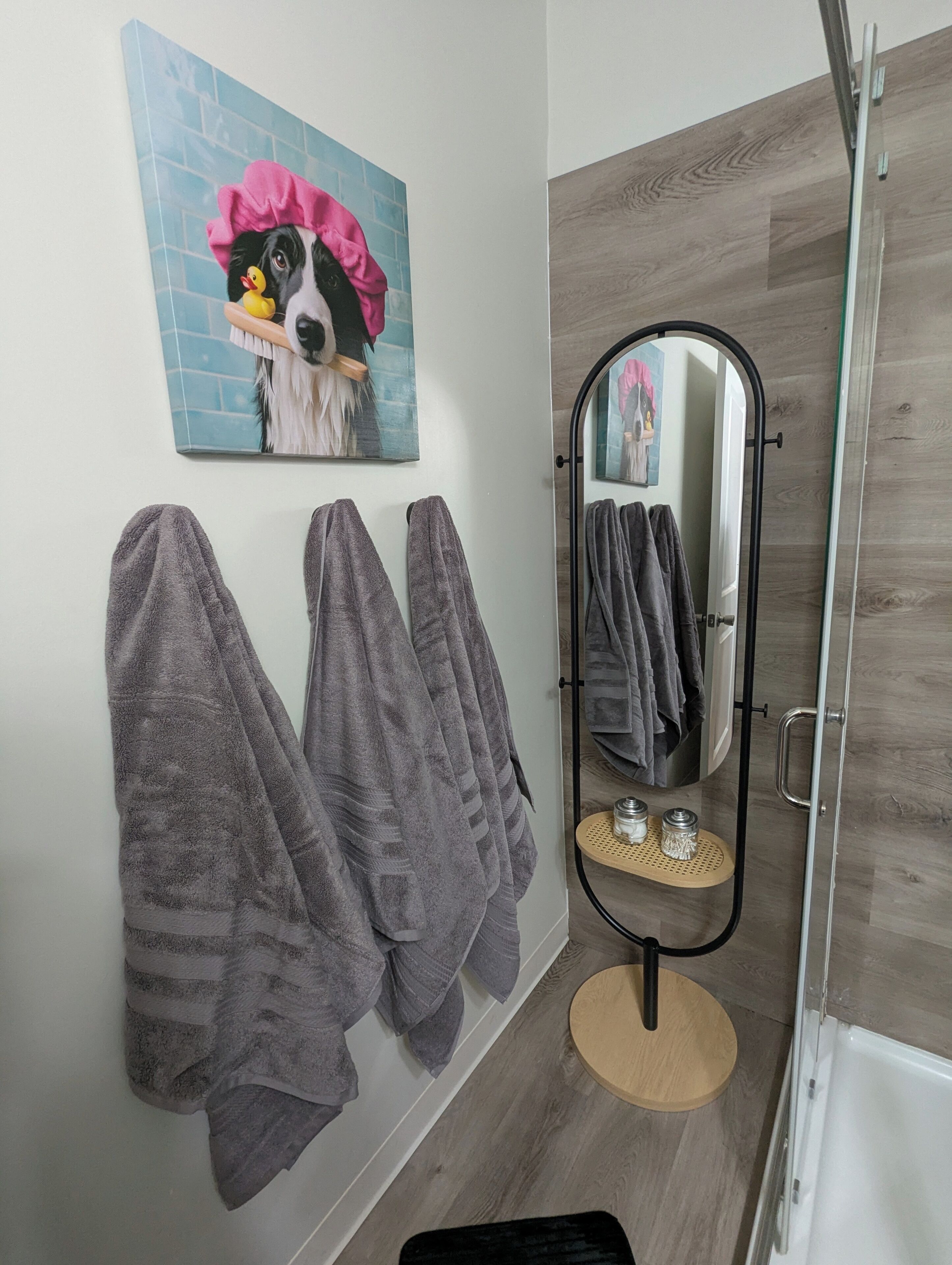A bathroom with a wooden floor, a mirror with a black frame, and a towel rack with three towels.