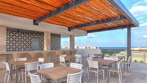 An outdoor patio area with multiple tables and chairs, a patterned wall, and a view of the ocean. - Beach Club Access & Rooftop Pool in La Amada (Cancún)