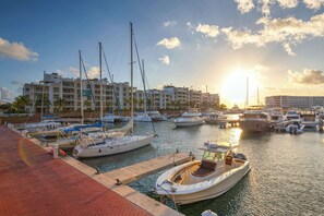 A marina with boats docked, a waterfront promenade, and a building complex in the background.