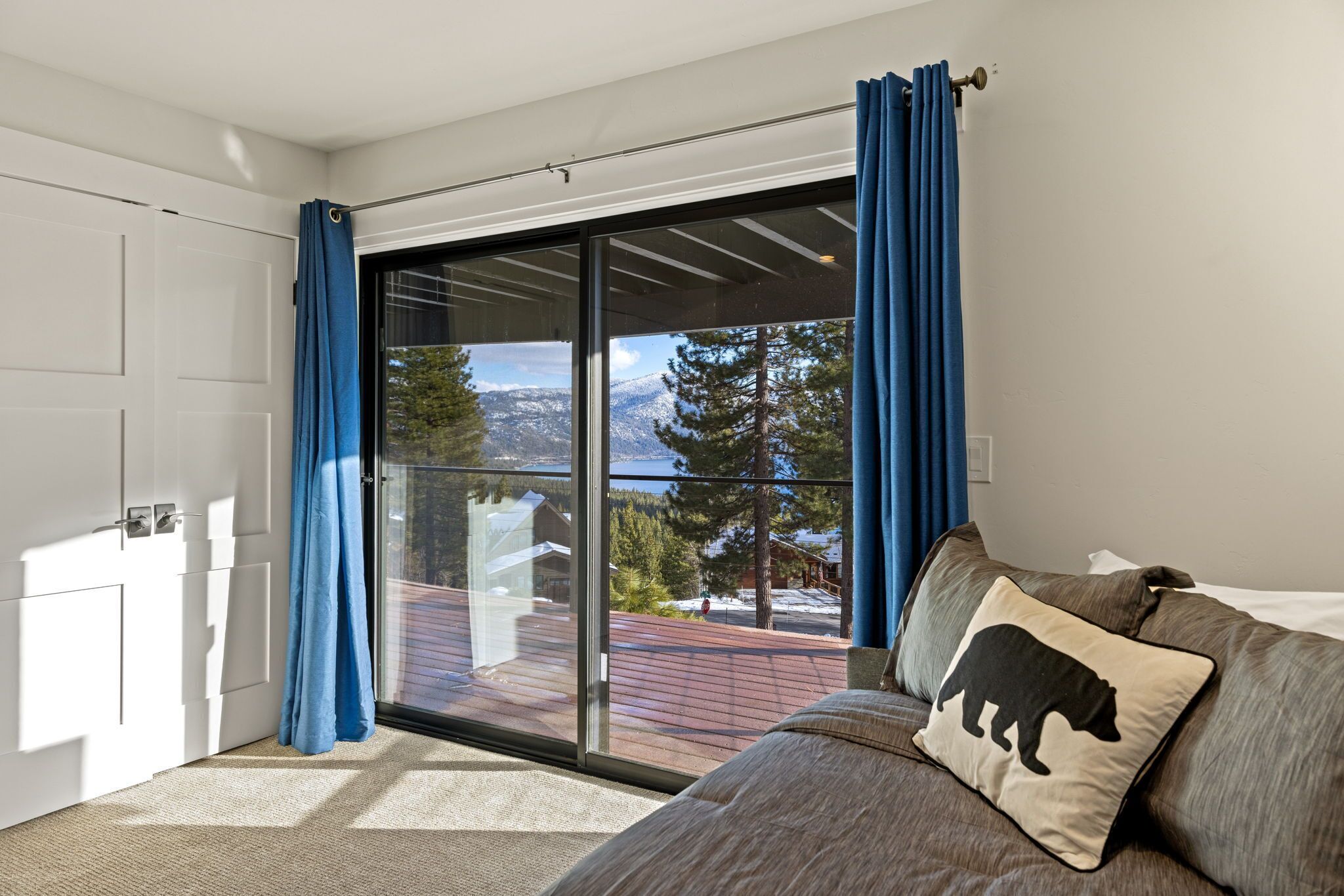 A room with a sliding glass door leading to a deck, a sofa with a bear pillow, and a view of a snowy landscape.