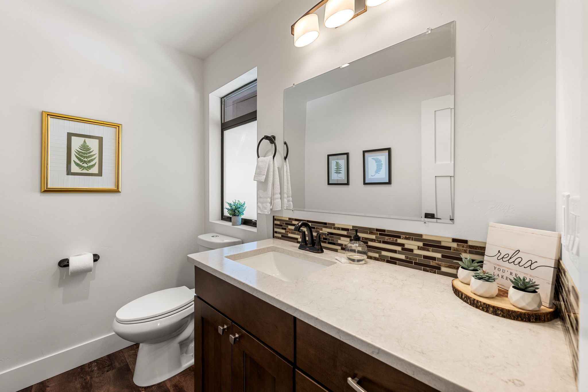 Un baño moderno con un inodoro blanco, un lavabo con encimera color beige y un espejo grande sobre el mueble. Hay cuadros enmarcados en la pared y una planta pequeña en el mueble.