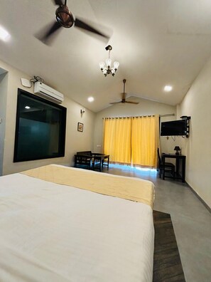 A hotel room with a large bed, a ceiling fan, an air conditioning unit, a TV mounted on the wall, a desk with a chair, and a window with yellow curtains. - MOUNTAIN BREEZE RESORT (Mahabaleshwar)