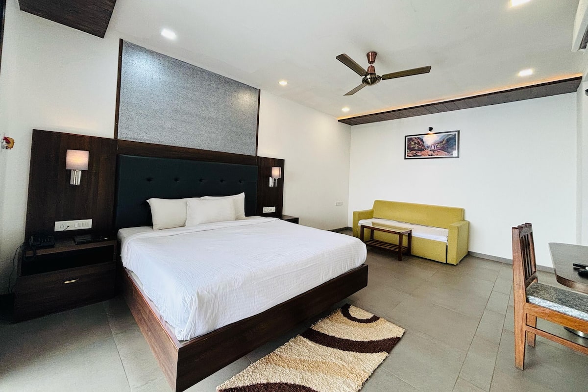 A modern hotel room with a large bed, a ceiling fan, a small sofa, and a dining table.