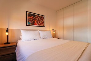 Un lit bien fait, avec du linge de lit blanc, un tableau encadré au mur et une armoire aux portes coulissantes.