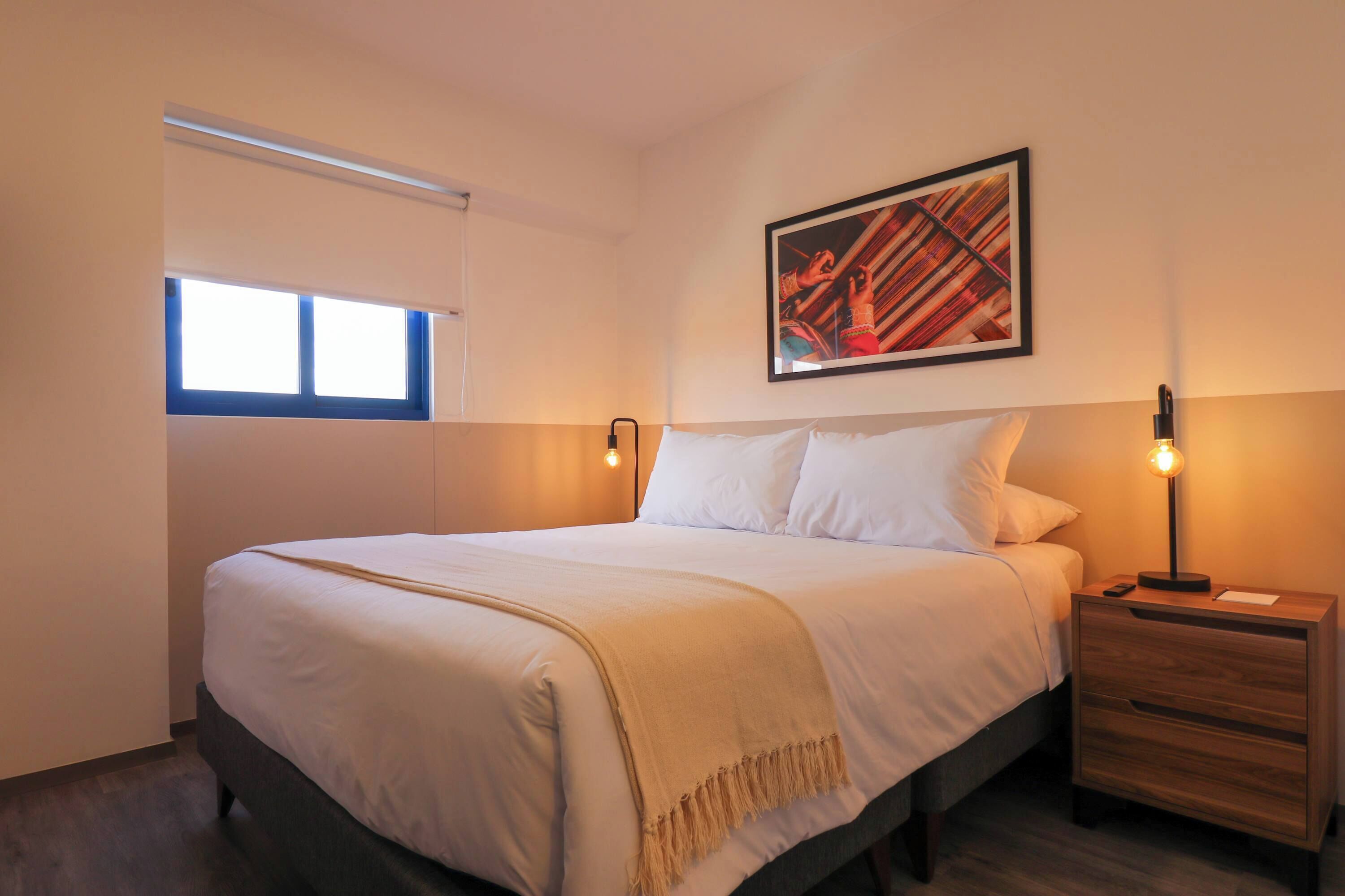 Una cama bien hecha con edredón blanco y manta color beige, una mesita de noche con lámpara y un cuadro abstracto enmarcado en la pared.