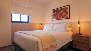 Un lit bien fait, recouvert d’une couette blanche et d’un plaid beige, une table de chevet avec une lampe, et un tableau abstrait encadré accroché au mur.
