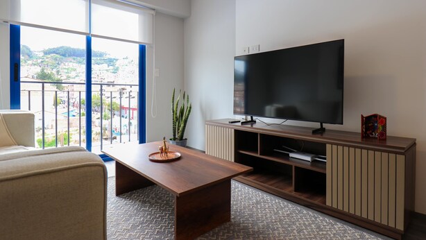 A modern living room with a large TV, a wooden coffee table, and a view of a cityscape through large windows. - w* | Modern 2BR in Historic Cusco w/ Balcony (Cusco)