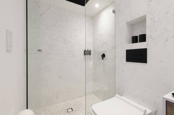 Une salle de bain moderne dotée d’une douche à parois de verre, de murs en marbre blanc et d’une étagère intégrée pour les articles de toilette.