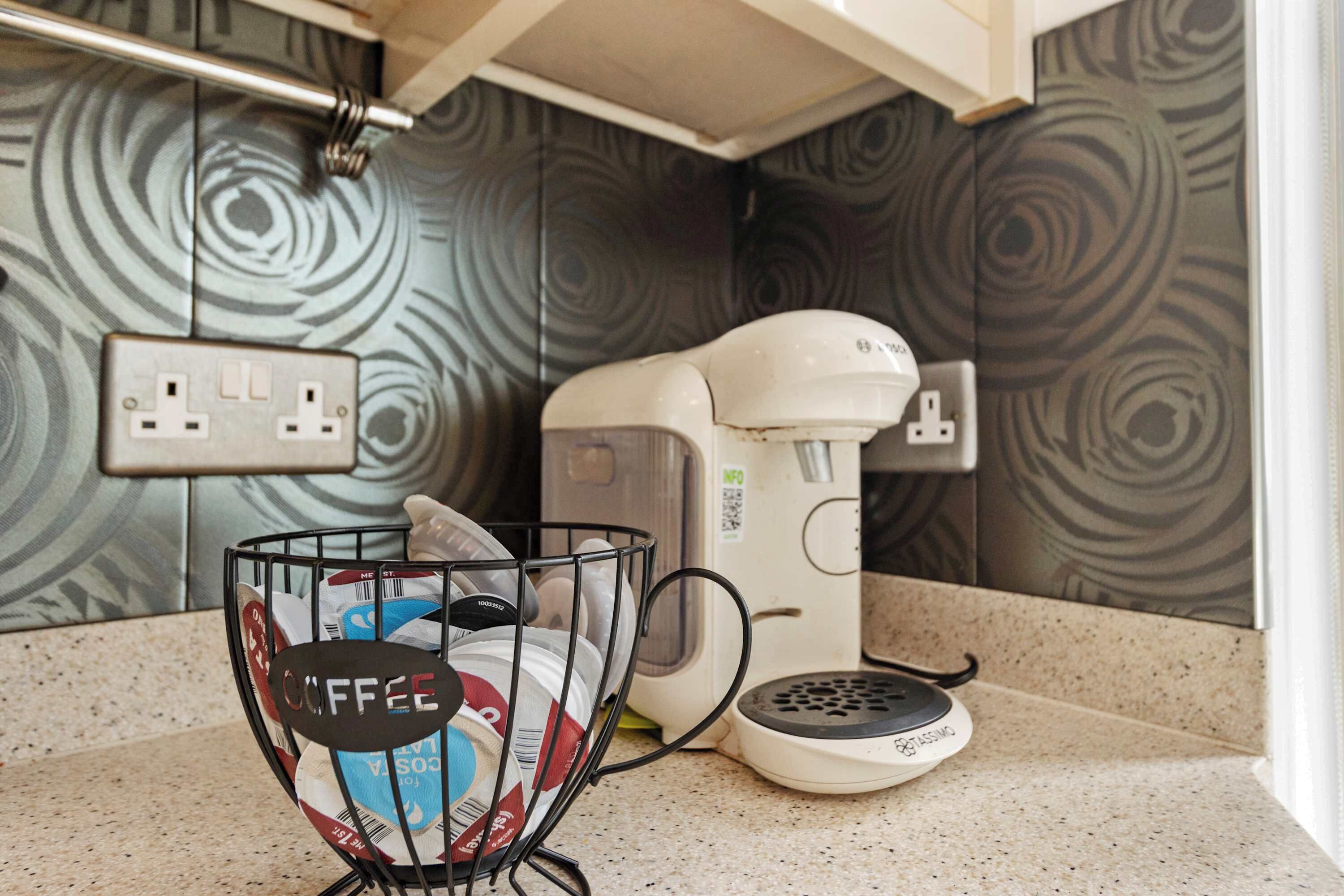 A kitchen with a coffee maker, a power outlet, and a coffee cup holder with a coffee bag.