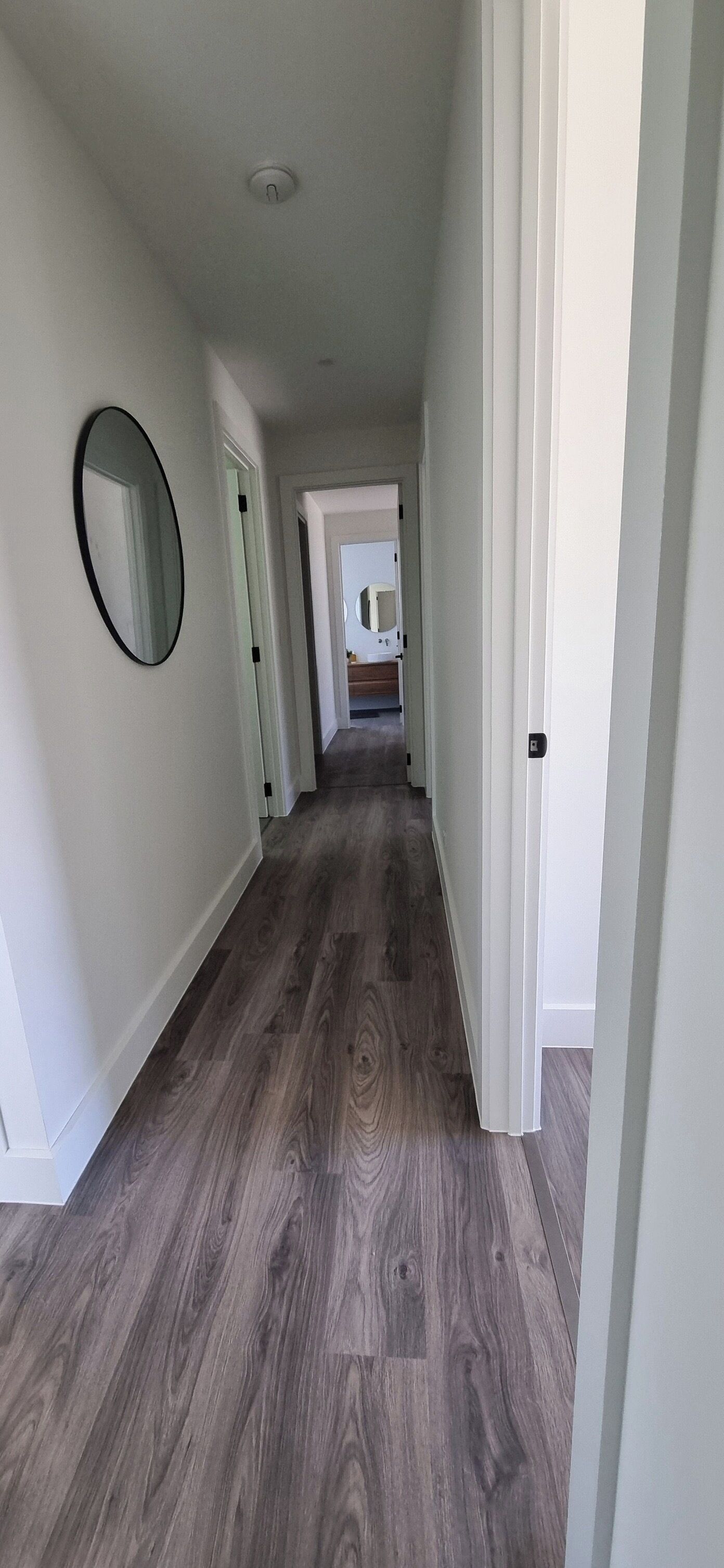 Un couloir doté d’un plancher en bois, de murs blancs et d’un miroir rond sur le côté gauche.